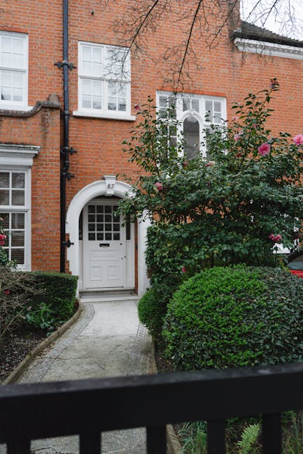 A view of a charming red brick townhouse with a white front door set within an arched brick surround, accessed via a narrow, curved paved pathway. To the right of the entrance, a lush, well-maintained