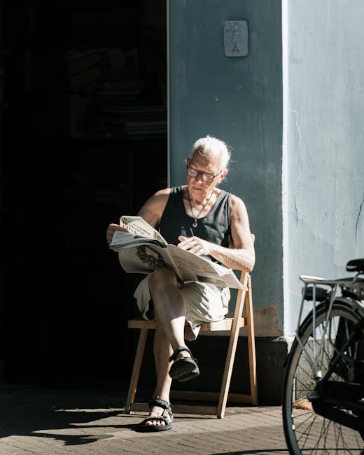 An elderly woman with short gray hair and glasses is seated on a small wooden stool outside a building with a light gray wall. She is wearing a sleeveless black top, beige shorts, and black sandals, a